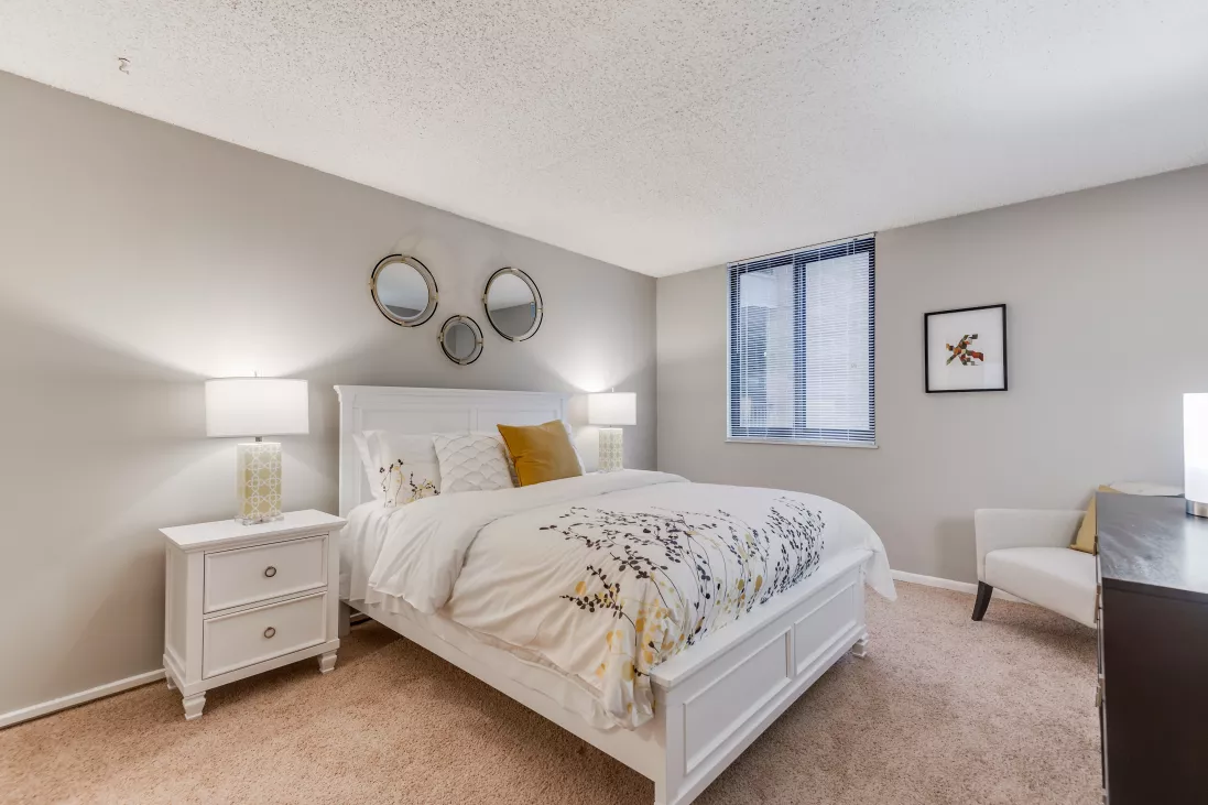 Bright bedroom with a white bed, yellow and gray floral bedding, two nightstands with lamps, and decorative mirrors.
