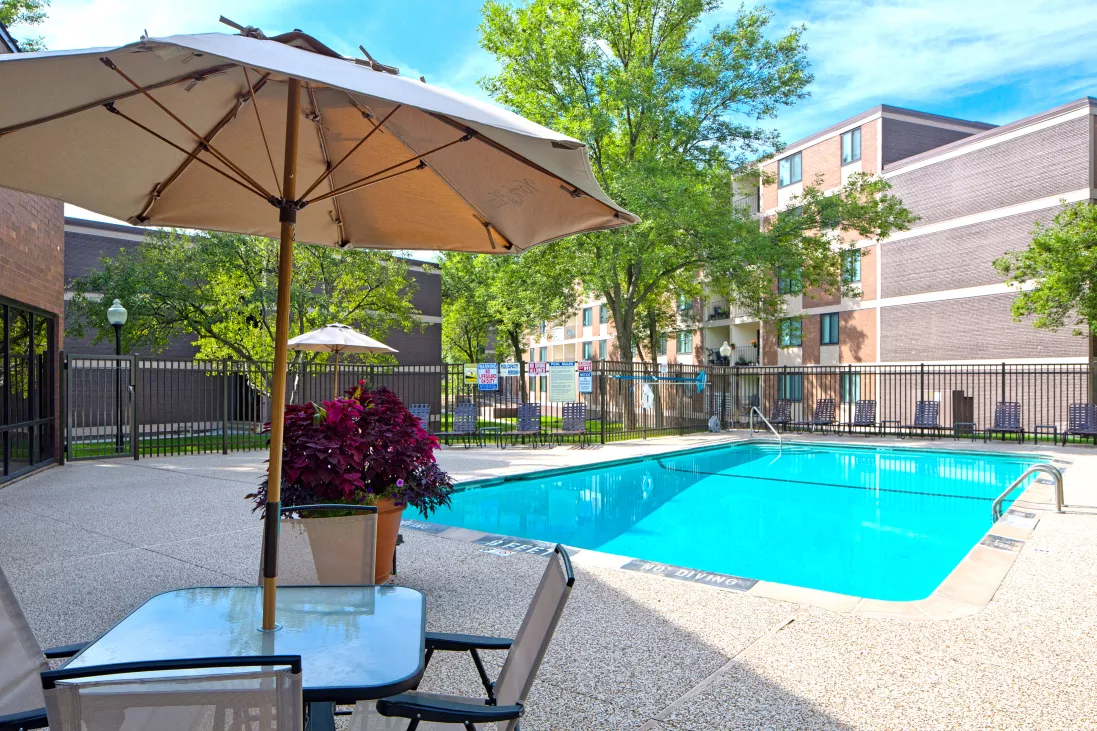 Sunny outdoor swimming pool with a patio table, chairs, and beige umbrella. Apartment buildings and green trees in the background.
